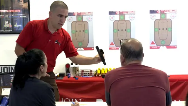 An instructor demonstrating a pistol to students in the Miami Shooters Club classroom, with dummy rounds on the table and laser targets on the wall