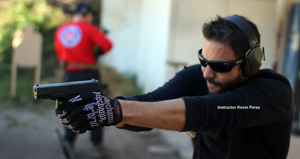 Instructor Kevin Perez shooting a pistol with a two-handed grip while another student shoots downrange